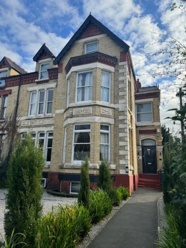Victorian-style house with bay windows, surrounded by shrubs and a cloudy sky.